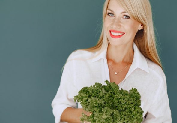 Cheerful woman with blonde hair holding fresh lettuce, promoting healthy living in a studio setting.