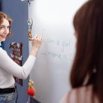 Smiling teacher writing on a whiteboard in a lively classroom setting with students.
